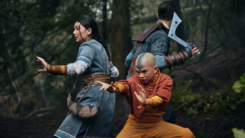 Three cosplayers in a forest scene portraying characters from Avatar: The Last Airbender, featuring Aang in the foreground with Katara and Sokka in action poses.