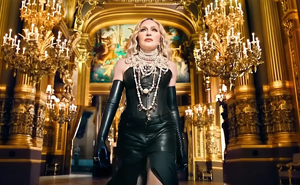 Elegant woman in black leather outfit and layered pearl necklaces standing in opulent room with golden chandeliers and classic paintings