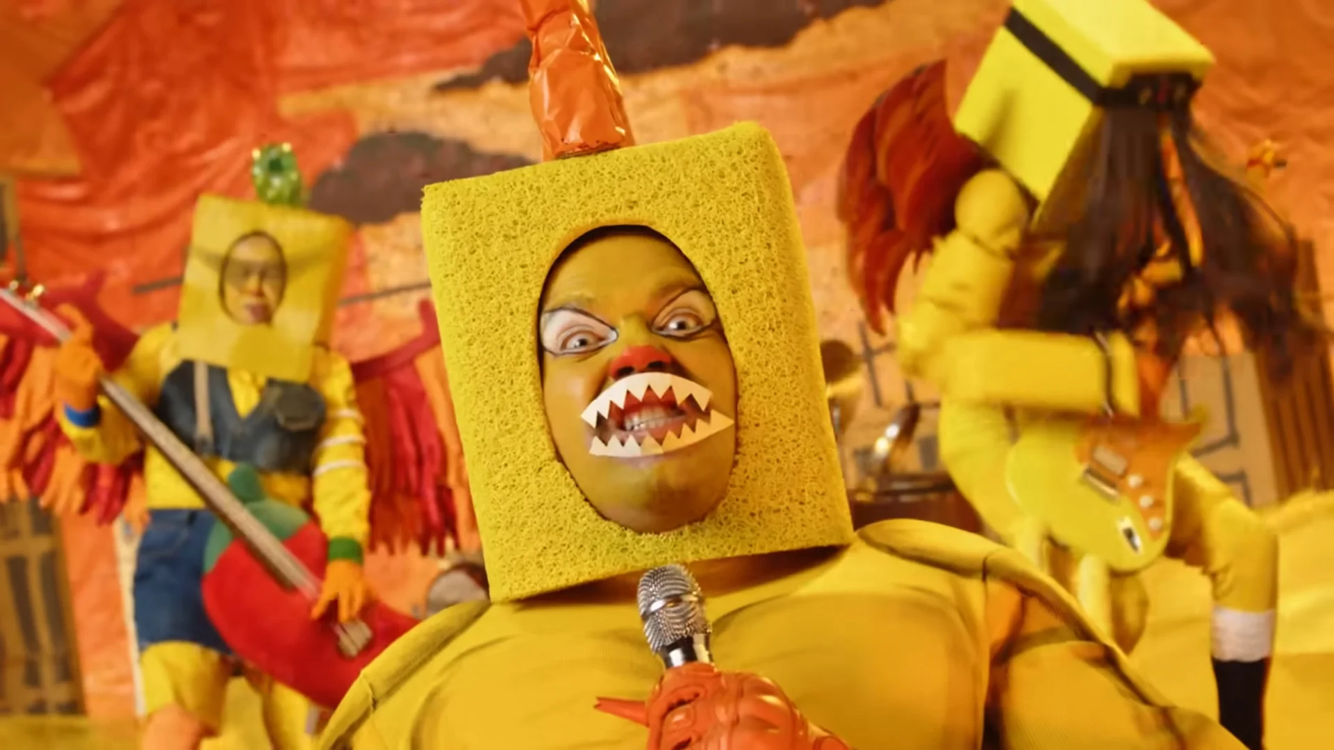 Person in sponge costume performing with microphone on stage with band members in quirky outfits.