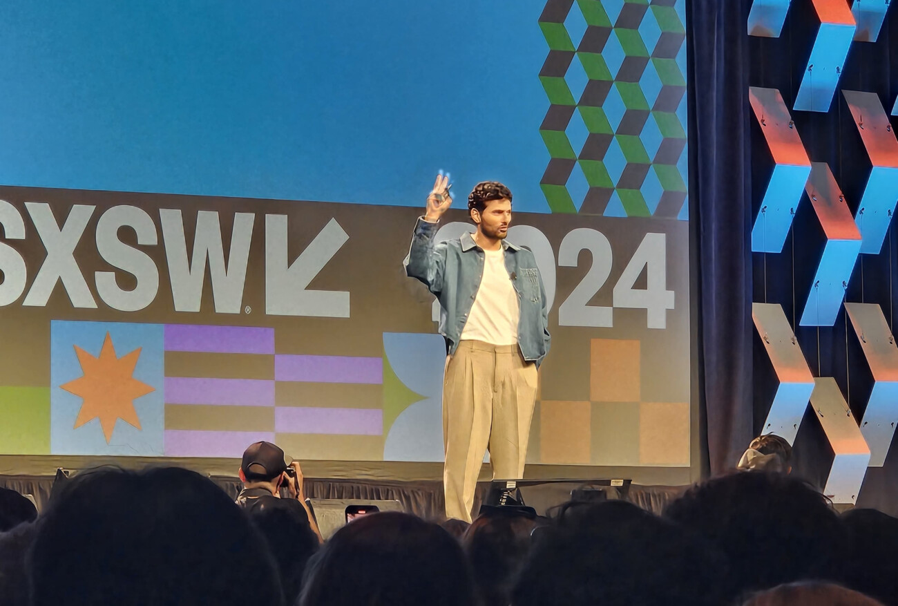 Speaker giving peace sign on stage at SXSW conference with audience and colorful backdrop