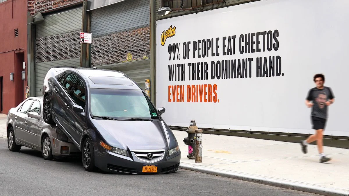 Car parked on top of another car on urban street next to a large billboard with text about people eating snacks with dominant hand.