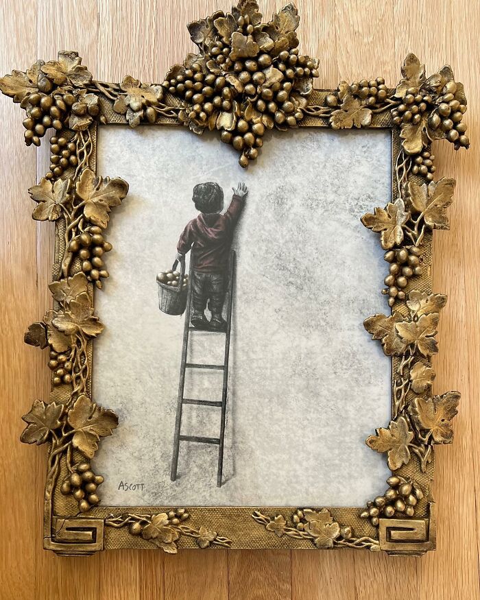 Artwork of a person on a ladder painting within an ornate golden grape cluster frame on a wooden surface.