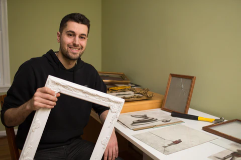 Young man smiling and holding a white picture frame in a workshop with framing tools in background.