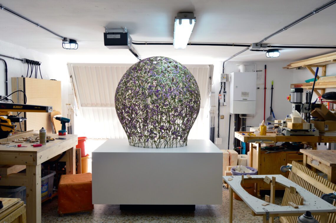 Sculpture installation of a large egg-shaped artwork adorned with purple flowers in a well-equipped garage workshop.