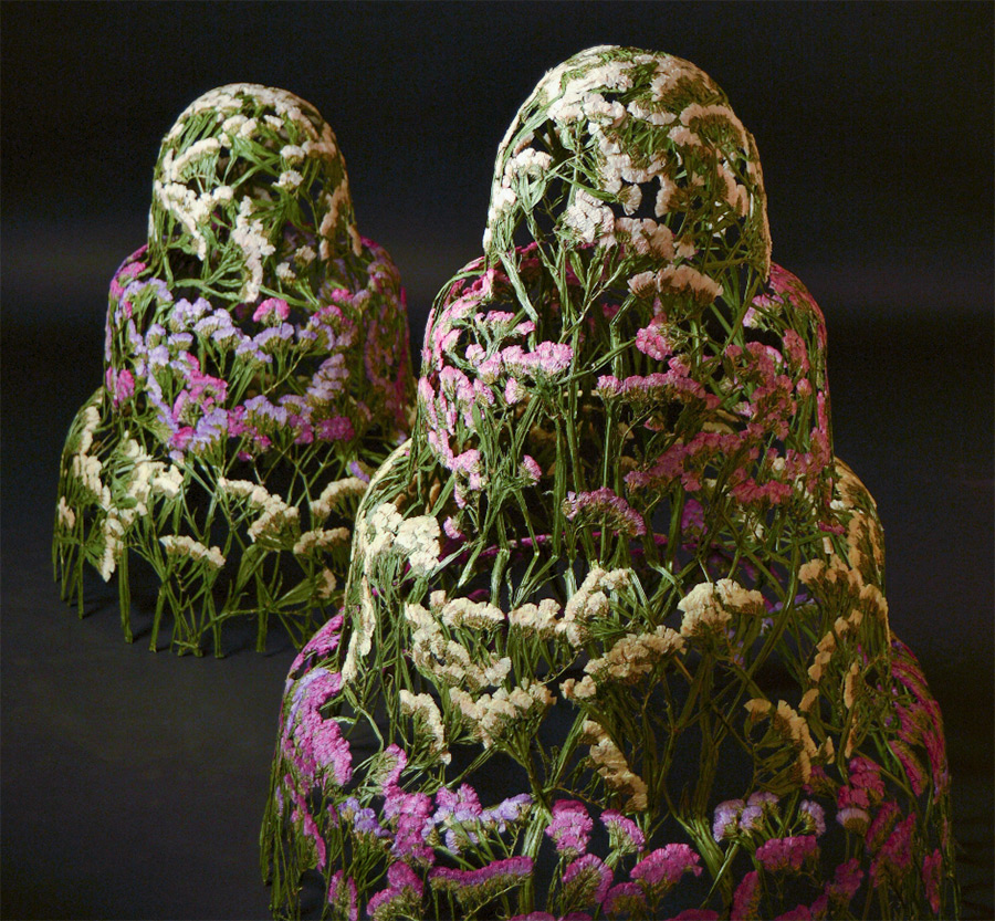 Artistic floral sculpture display with pink and white flowers arranged on two human-like figures against a dark background.