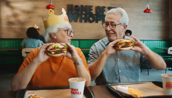 Senior couple enjoying burgers at a fast-food restaurant with one wearing a paper crown.