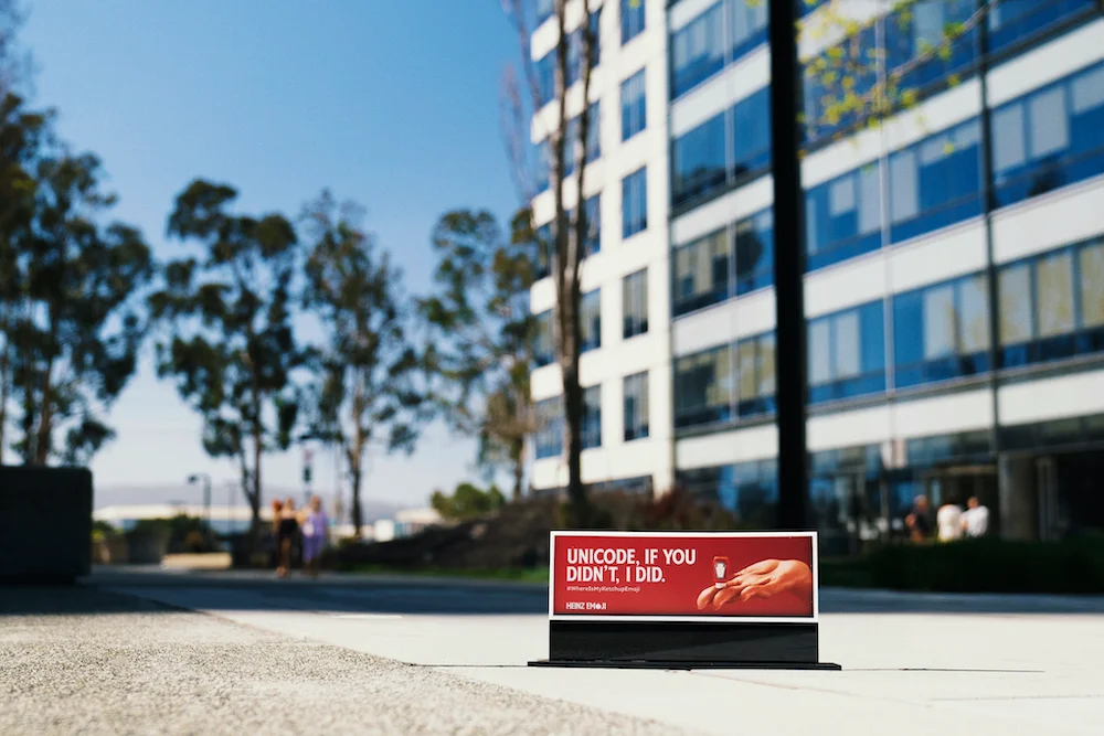 Low-angle view of an advertisement board on city sidewalk with focus on text "UNICODE, IF YOU DIDN'T, I DID" and blurred modern building and people in the background
