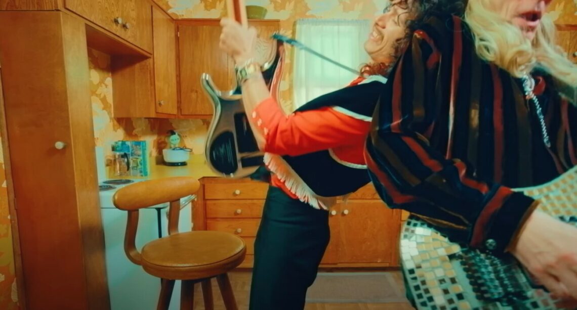 Two people playing air guitar enthusiastically in a vintage kitchen with retro wallpaper. Joe Satriani e Steve Vai