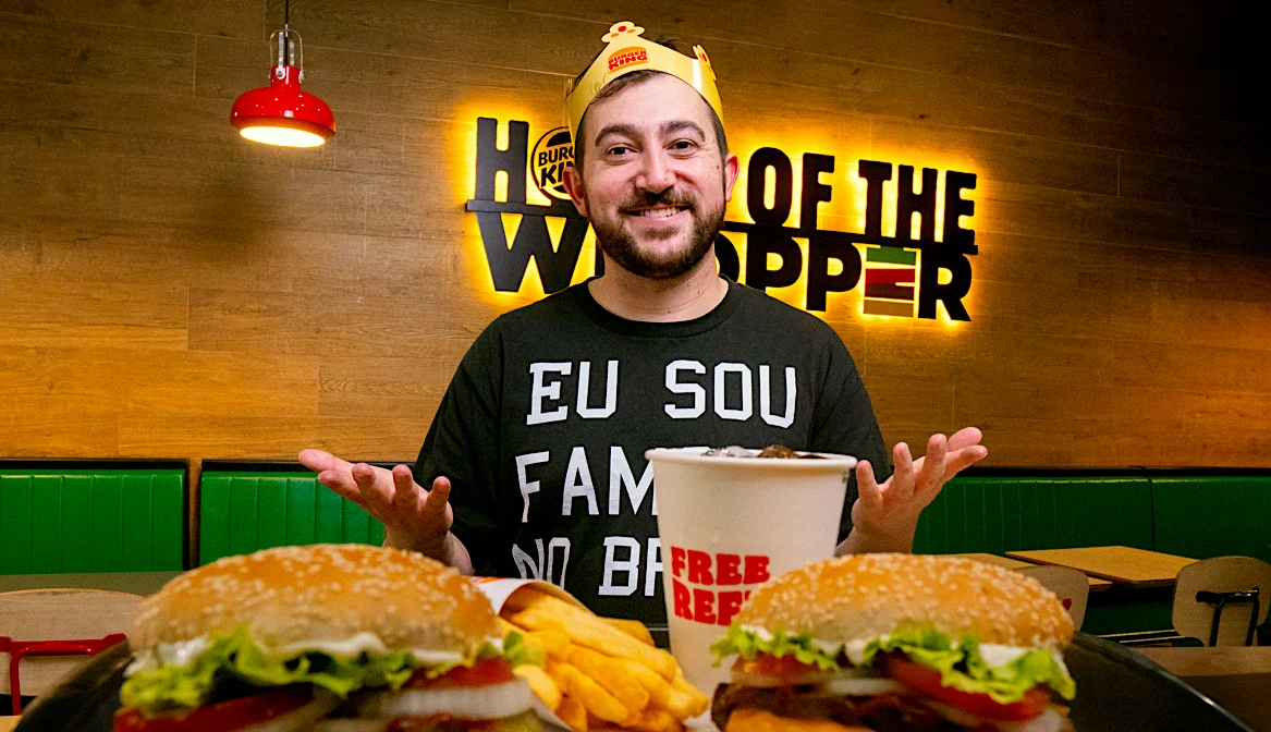 Man in Burger King crown smiling with Whopper burgers and soda in a Burger King restaurant.