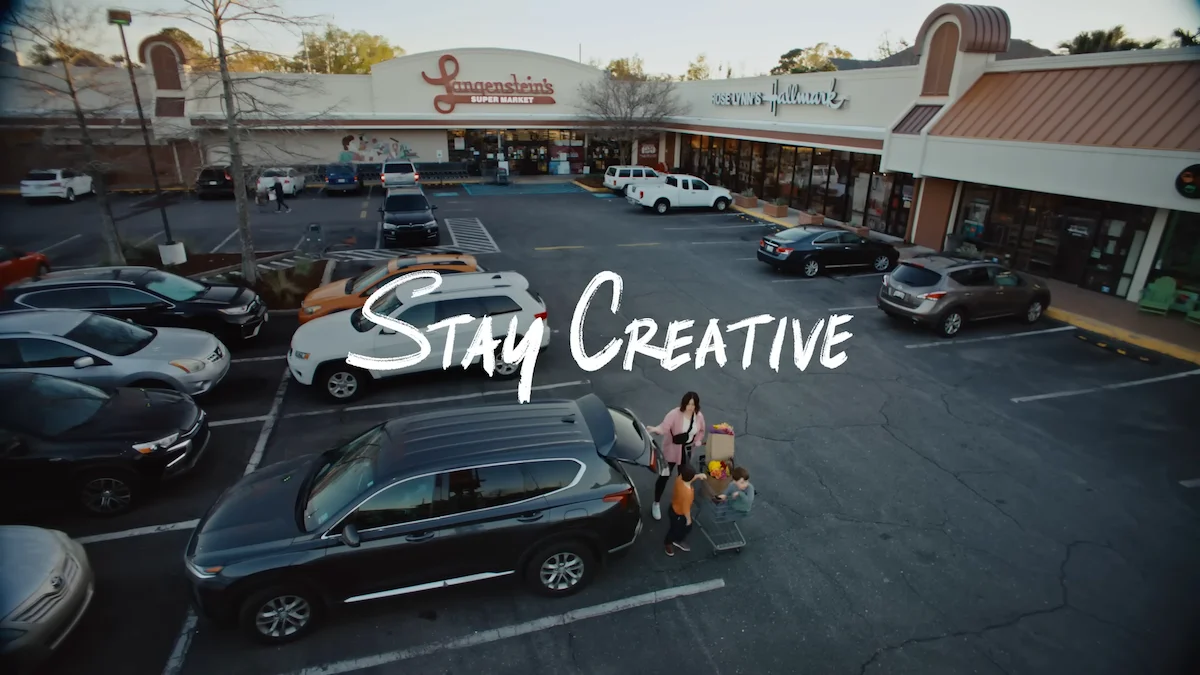 Outdoor shopping center parking lot with cars and a family with a shopping cart under the "Stay Creative" text overlay.