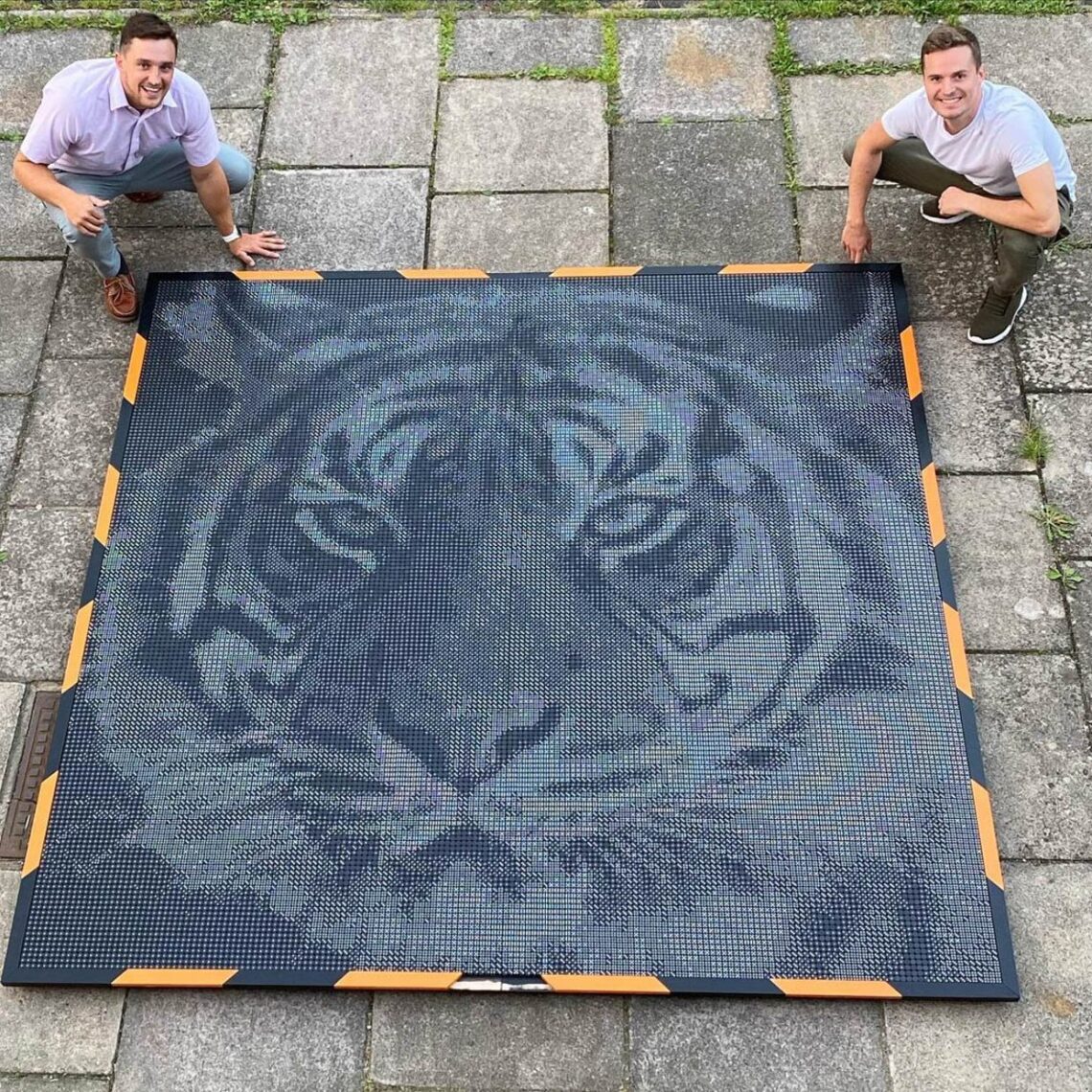 Two men proudly presenting a large tiger portrait made of dominoes on an outdoor patio.