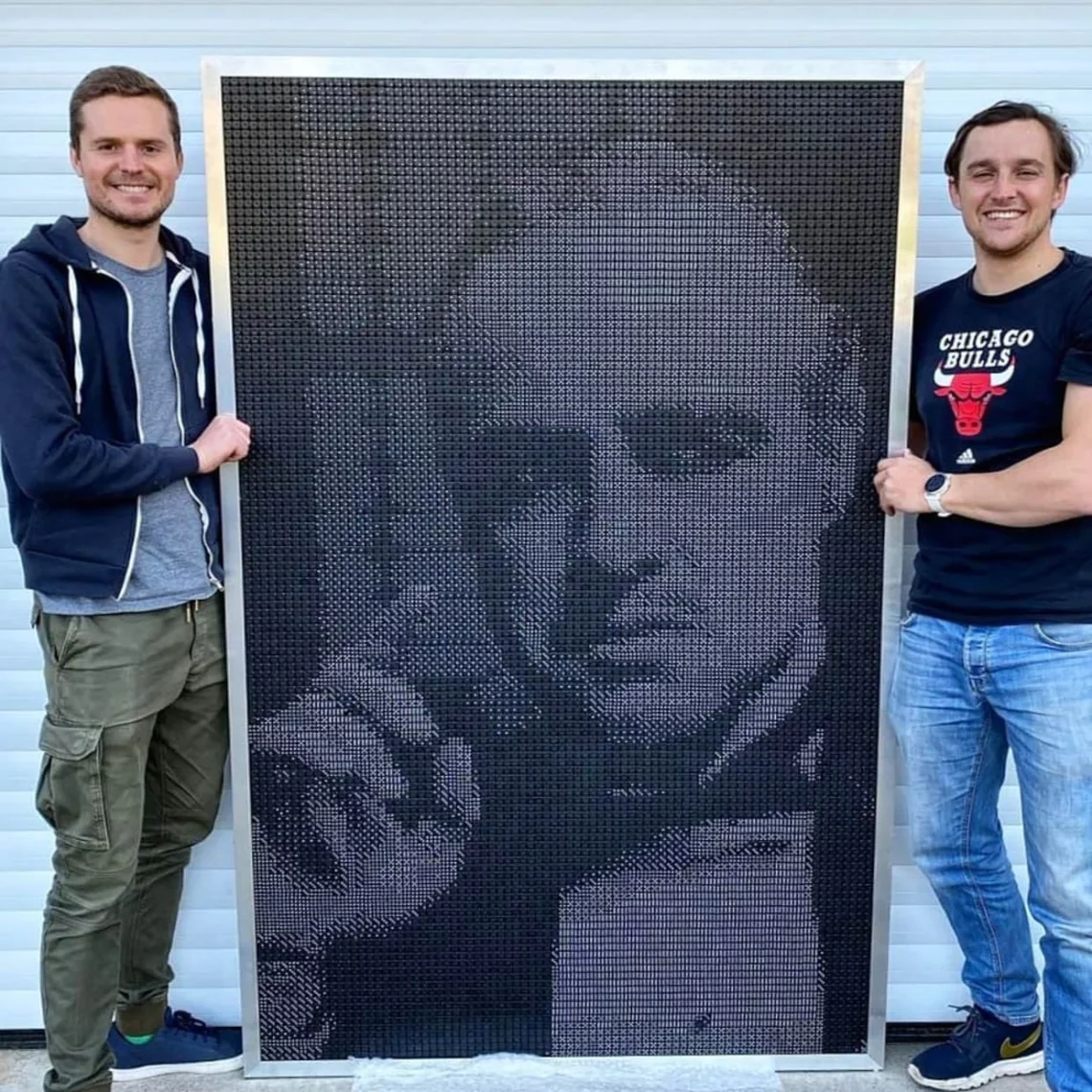 Two men standing beside a large monochrome mosaic portrait art piece.