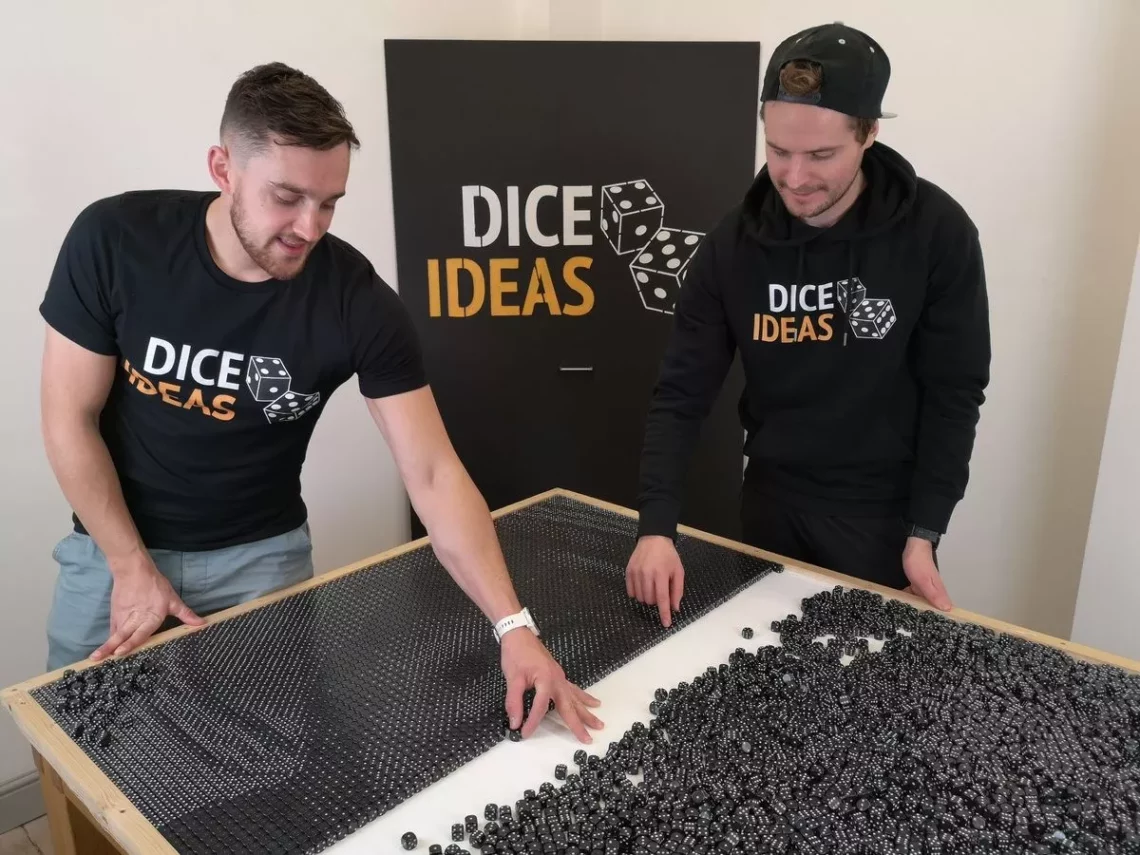 Two men assembling art installation with numerous black dice on a table in a workshop with DICE IDEAS branding