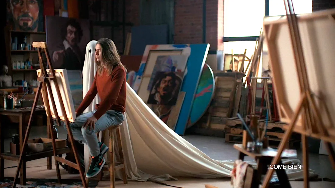Woman sitting on a stool in an art studio with paintings and a person under a sheet posing as a ghost.