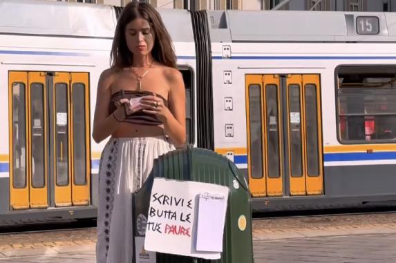 Woman checking phone by tram stop with a litter bin in foreground displaying a sign in Italian.