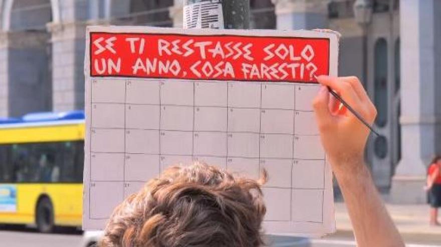 Person writing on a whiteboard with an Italian phrase asking "If you had only one year left, what would you do?" in a public setting with a city bus in the background.