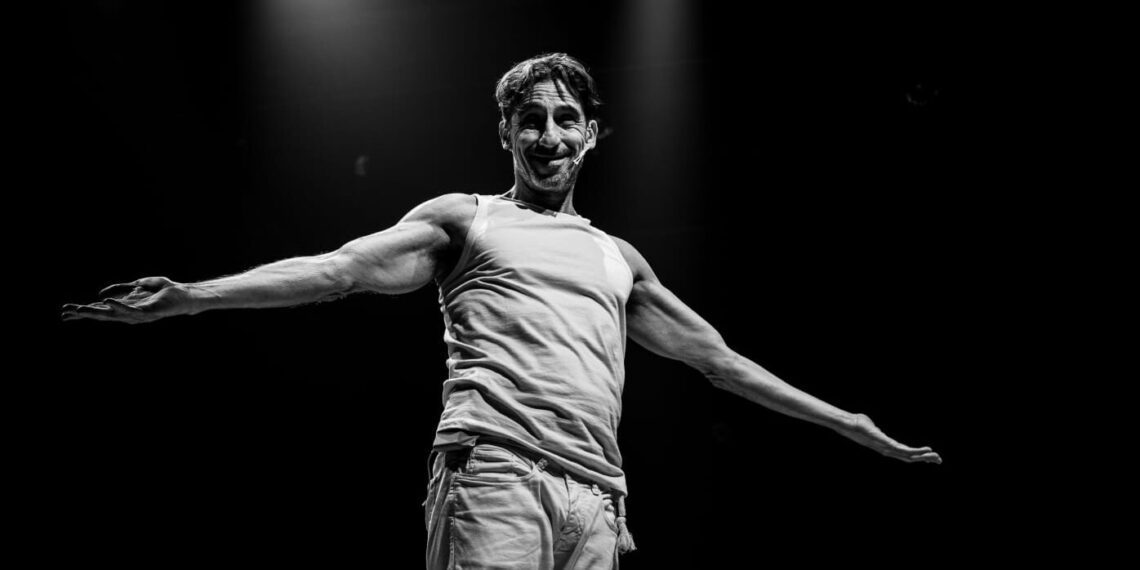Black and white photo of a smiling male performer on stage extending arms with a tank top and casual pants