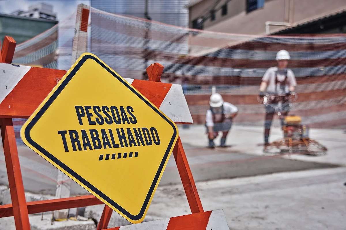 Yellow caution sign reading "PESSOAS TRABALHANDO" with blurred construction workers in background at urban worksite.