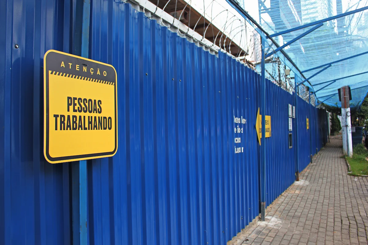 Yellow caution sign on blue construction site fencing with text 'Atenção Pessoas Trabalhando' indicating 'Attention People Working' in Portuguese.