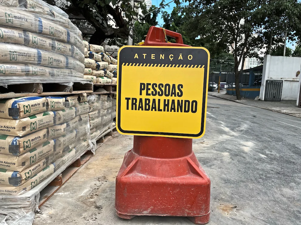 Yellow and black caution sign reading 'ATENÇÃO PESSOAS TRABALHANDO' in front of construction materials on a job site.