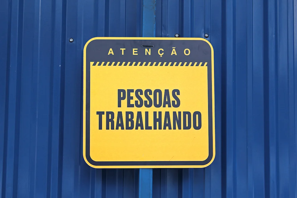 Yellow caution sign on blue corrugated surface with Portuguese text 'Atenção Pessoas Trabalhando' meaning 'Attention People Working'