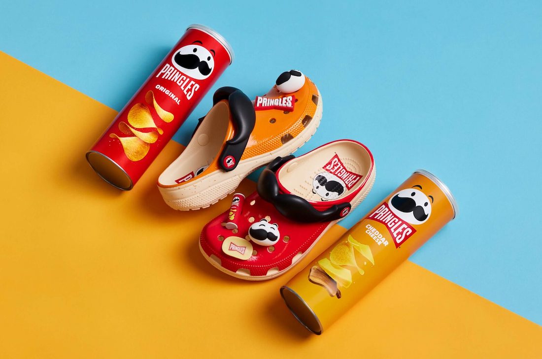 Pringles e Crocs. Pringles themed footwear collection featuring colorful clogs with iconic chip mascot alongside original and cheddar cheese flavor Pringles cans on a blue and yellow background.