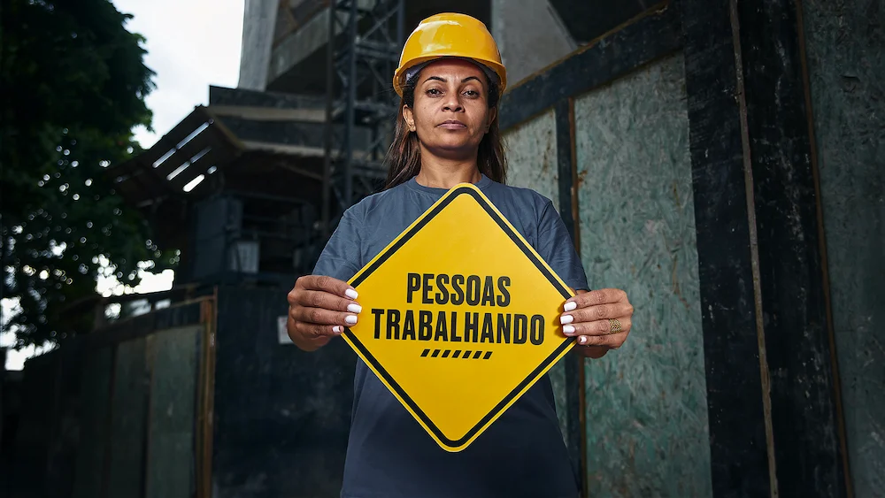 Construction worker in hard hat holding "PESSOAS TRABALHANDO" caution sign at building site