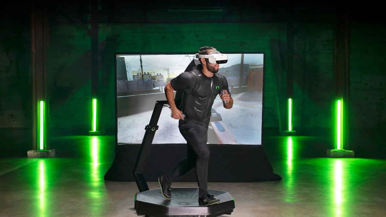 The Omni. Man using VR headset on omnidirectional treadmill for immersive gaming experience in a room with green lighting.