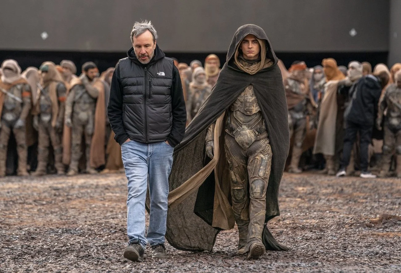 Director on movie set with actor in futuristic costume walking among extras in desert-like environment