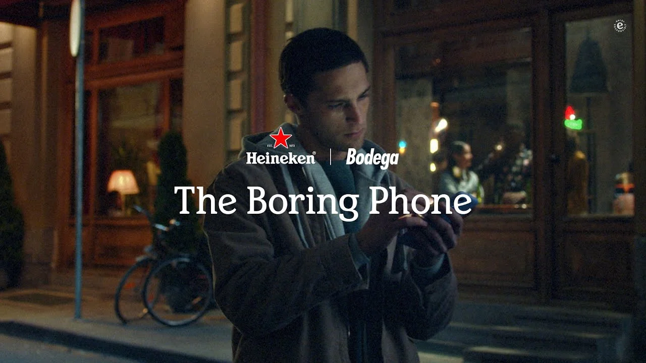 Man looking at his smartphone at night on a city street with the text "The Boring Phone" displayed alongside a Heineken logo and the word "Bodega".