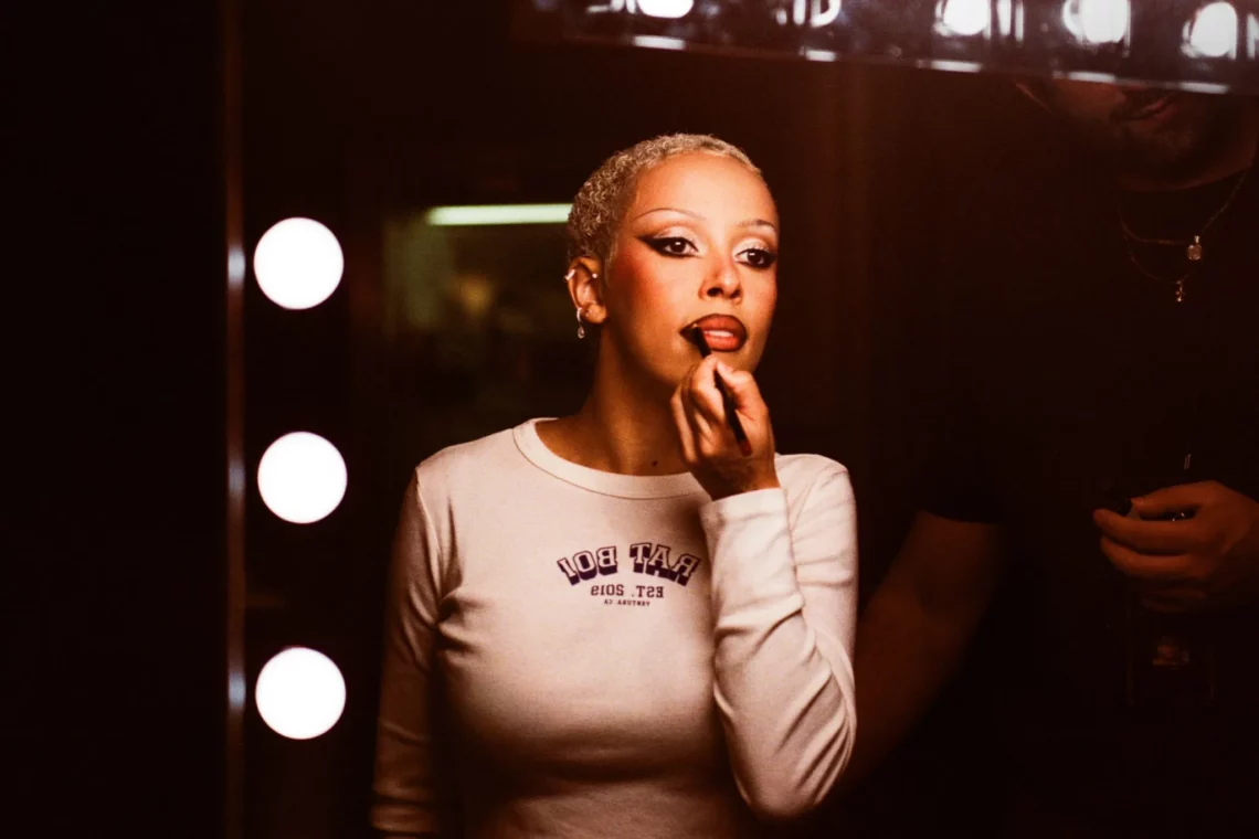 Woman with short platinum blonde hair applying makeup in a dressing room with illuminated vanity mirror lights.