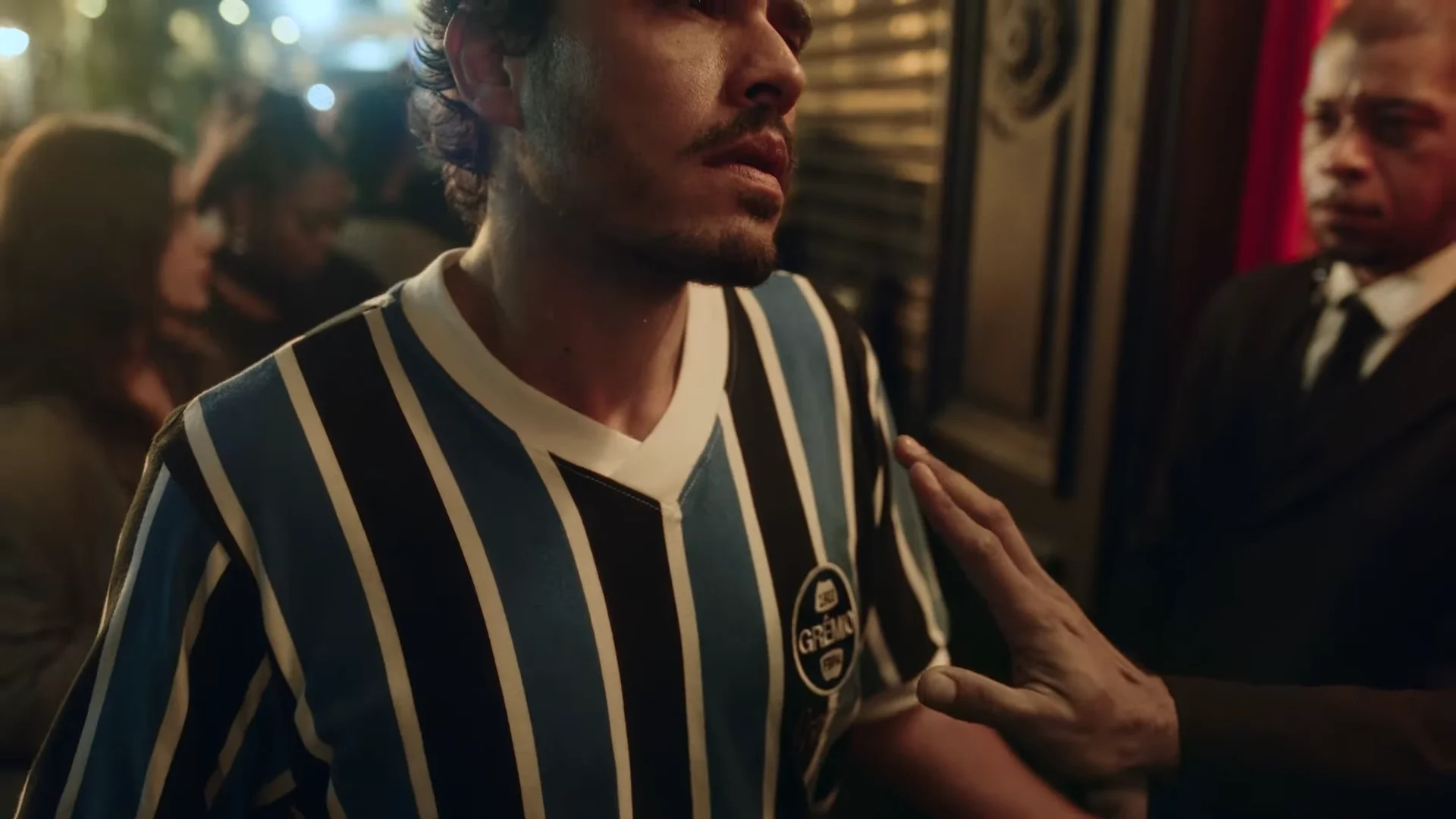 Alfaiataria Brahma: camisas de times na moda. Man in blue and white striped soccer jersey in a busy bar with a bouncer in the background.