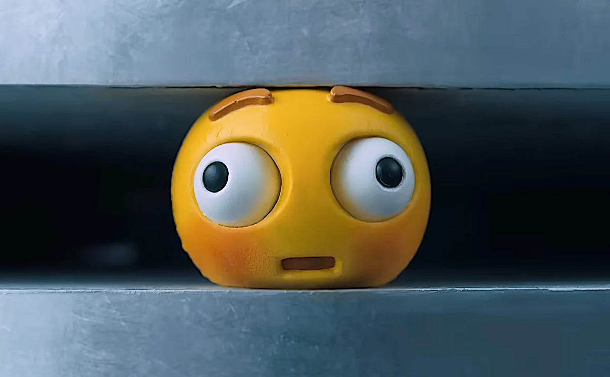 Yellow emoji stress ball with surprised expression peeking between gray metal blinds.