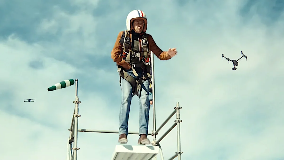 Man in helmet and harness standing on top of a high tower with a drone flying nearby against a cloudy sky background