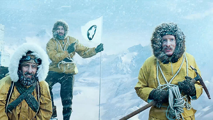 Three mountaineers in winter gear raising a flag on snowy peak.