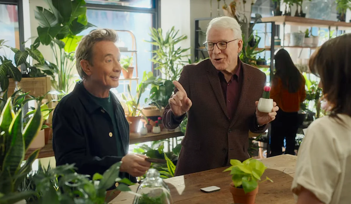 Two men having a conversation in a plant-filled indoor space, one holding a small cactus while gesturing to a woman across the table.