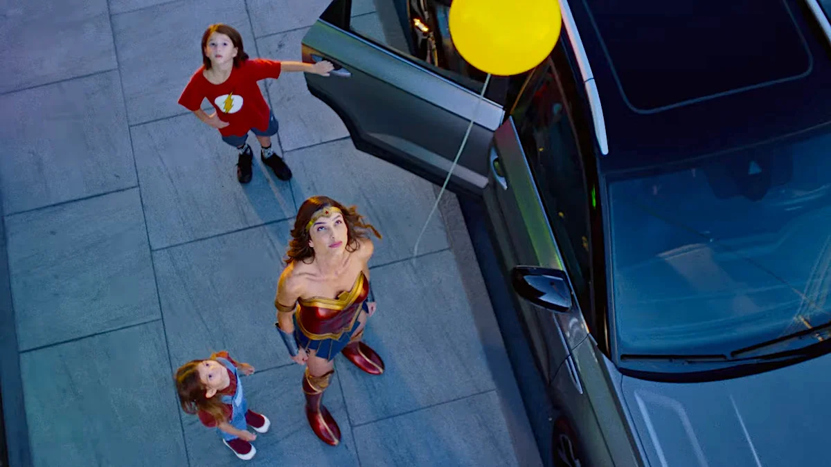 Woman dressed as Wonder Woman with children in superhero outfits standing by car with yellow balloon.