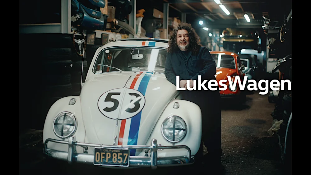 YourWagen. Man standing next to classic white Volkswagen Beetle with racing stripes in a vintage car garage.
