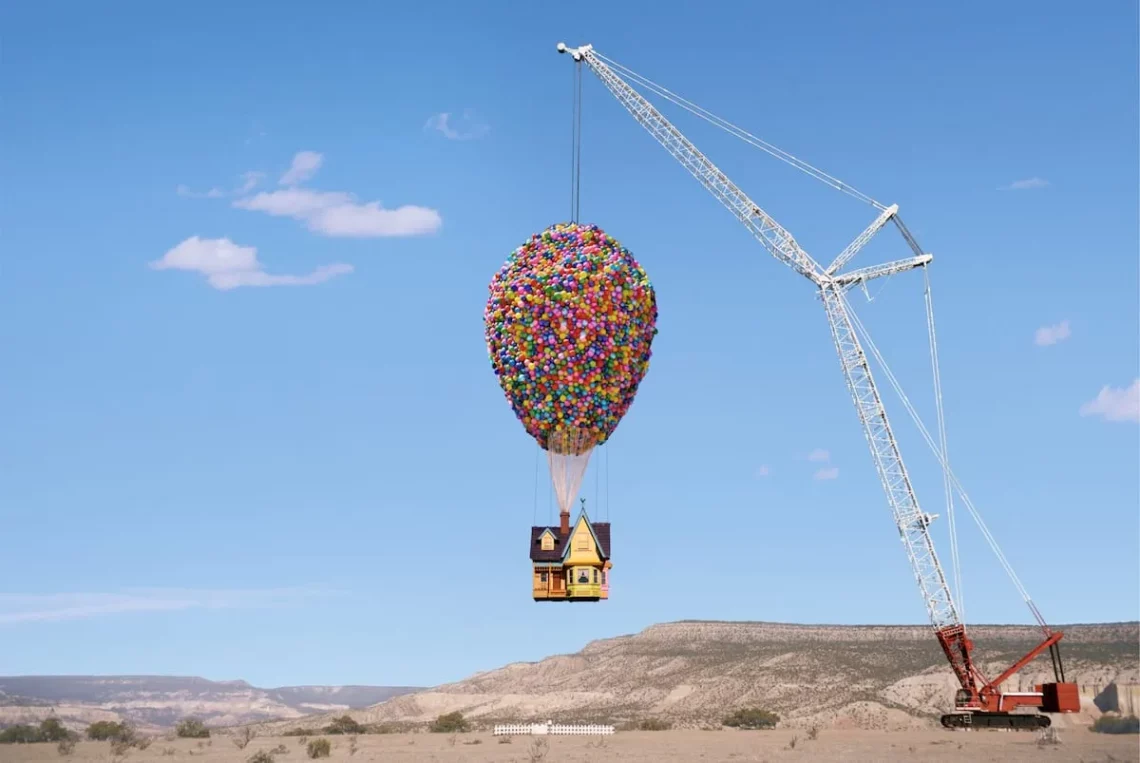 Colorful balloon-covered house lifted by crane resembling a hot air balloon in clear blue sky with desert landscape background.