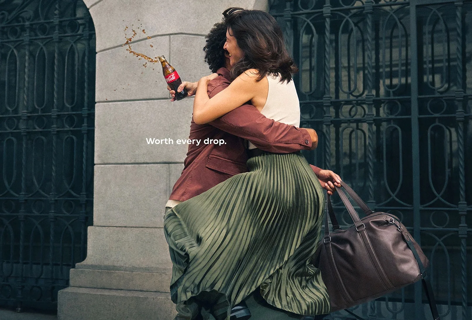 Woman hugging man while accidentally spilling Coca-Cola with tagline "Worth every drop" against ornate gate background.